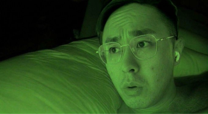 A man wearing glasses and an earpiece lies against a pillow in dim, green-tinted lighting, displaying a worried or skeptical expression. The eerie, surveillance-style atmosphere of the image humorously mirrors the cautionary tone of the post urging users to update their Phantom app before trading on the newly integrated BNB Chain.