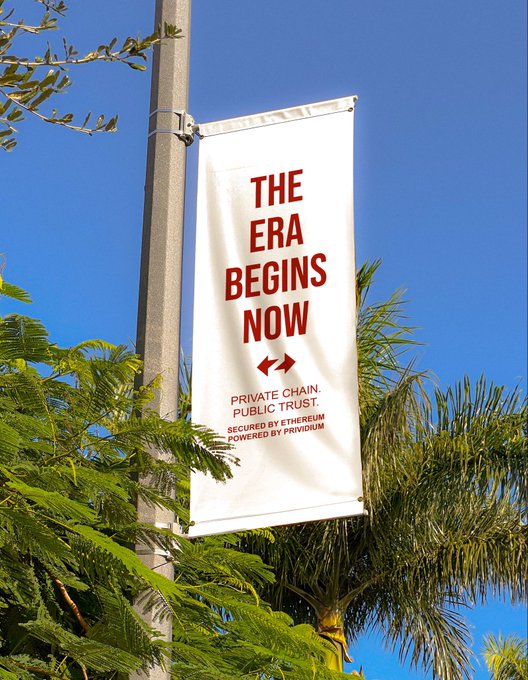 A vertical white banner mounted on a pole reads 'THE ERA BEGINS NOW' in red, with smaller text below stating 'PRIVATE CHAIN. PUBLIC TRUST. SECURED BY ETHEREUM POWERED BY PRIVIDIUM'.