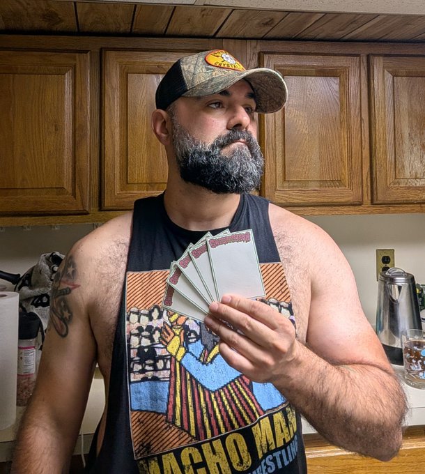 A bearded man wearing a 'Nacho Man' wrestling tank top and a camo baseball cap holds up a fan of stickers in a kitchen.