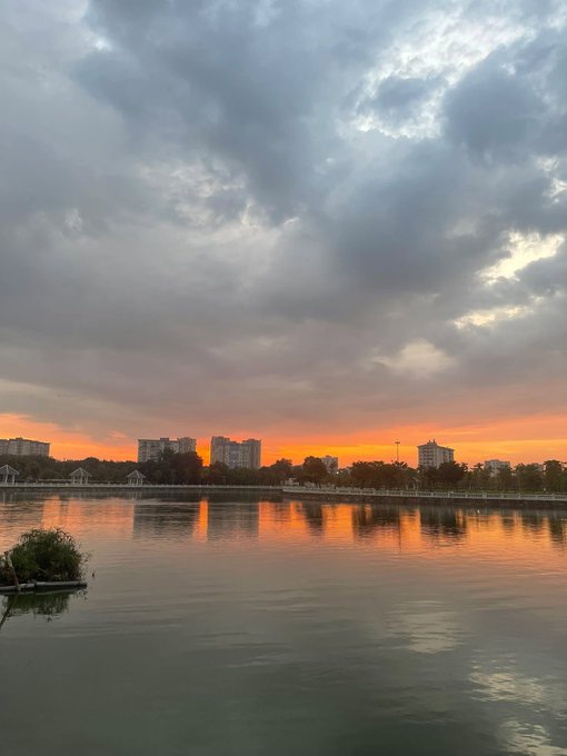 A scenic sunset over a calm lake with a cloudy sky and city buildings visible on the distant shoreline.