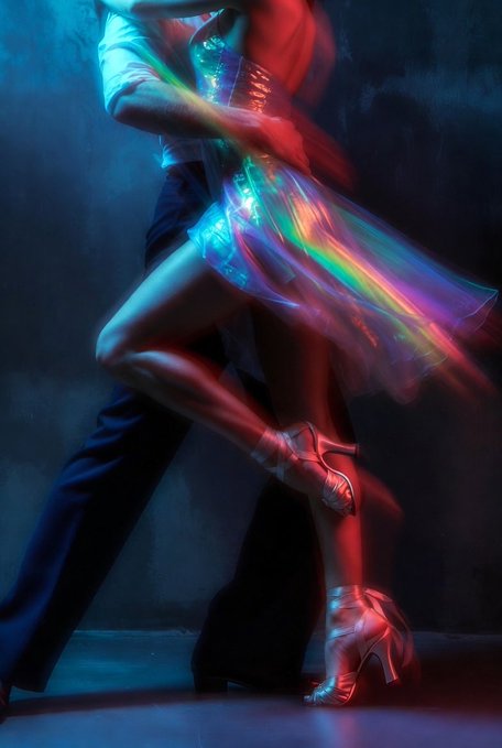 Artistic photograph of a dancing couple with motion blur, capturing the woman's iridescent flowing dress and red high heels under blue and red lighting.
