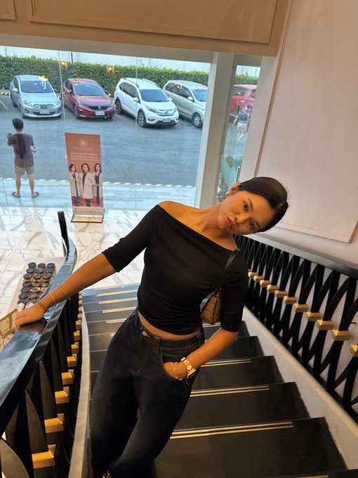 A woman wearing a black off-shoulder top and jeans poses on a staircase, with a parking lot visible through the window behind her.