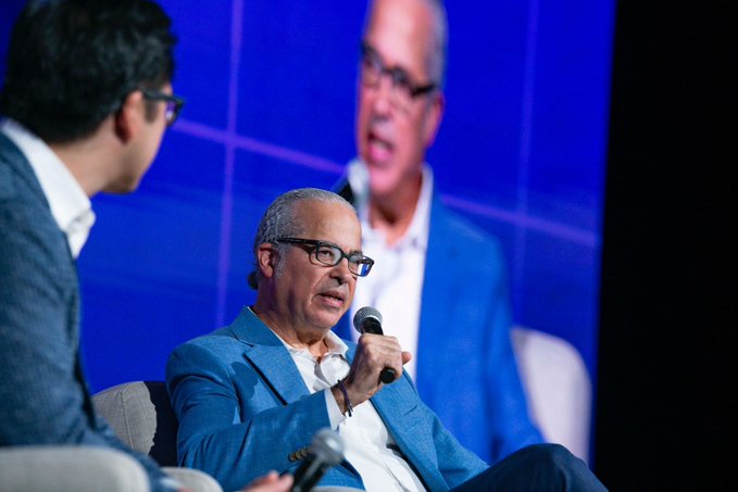 The image shows a man wearing glasses sitting down while another man stands behind him holding a microphone. The two men appear to be engaged in conversation or speaking about Bitcoin futures and Ether futures, as indicated by the text in the image.