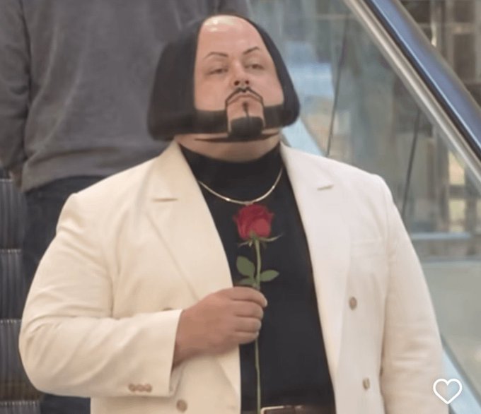 A person wearing a black turtleneck and a white blazer holds a red rose, with their head digitally altered to appear as a stylized, cartoonish mask with a goatee.