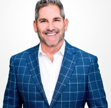 The image features a man wearing a blue plaid suit, smiling at the camera with his hands placed behind his back. The background of the photo is white, which contrasts with the man's dark suit. There are no other objects or texts present in the image.
