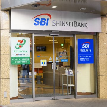 The image shows a bank building with a large glass entrance, displaying the name "SBI Shinsei Bank" above the door. The bank offers Bitcoin rewards for its customers, as indicated by the text provided in the image.