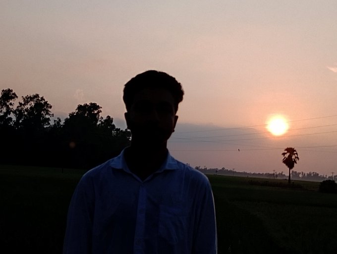A silhouetted photo of a person in a light blue shirt standing outdoors during sunset, with a tree-lined horizon and the sun low in the background.