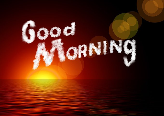 A scenic sunrise over the ocean with the text 'Good Morning' written in cloud-like letters, accompanied by lens flare effects.