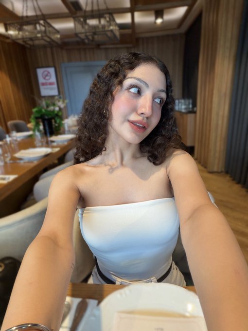 A young woman with curly hair in a white strapless top takes a selfie at an upscale restaurant, looking off to the side with a cheerful expression that mirrors the upbeat "life is good" sentiment.