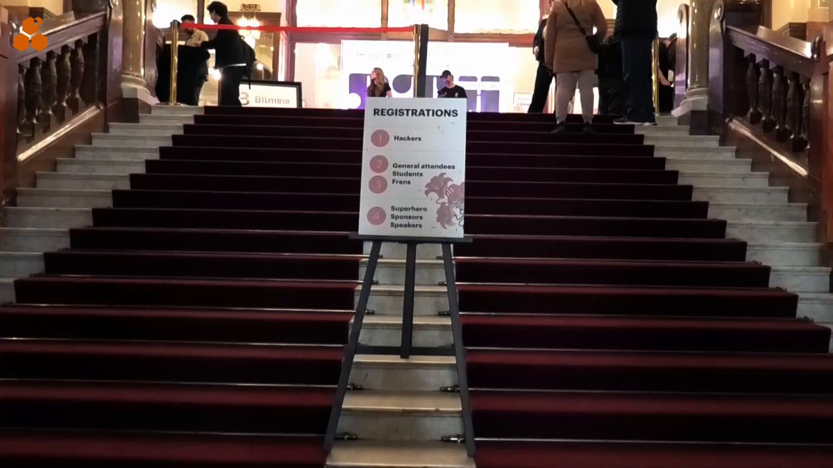 The image shows a group of people standing near stairs, with some of them looking down into an office or conference room. There are two signs posted on the steps that provide information about registration for a hackathon event. The first sign is located at the top of the stairs and reads "Registration". The second sign is placed lower down the stairs and says "Hackathons". A person can be seen in the background, possibly attending to one of these events or providing assistance.