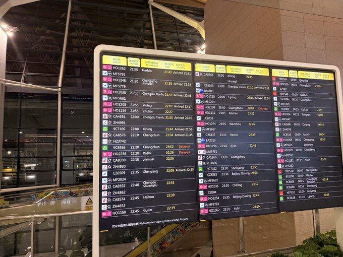 A large flight information display board at Beijing Capital International Airport (PEK) showing numerous domestic flight statuses, including arrivals from Harbin, Chongqing, Xining, and Shenzhen, with several marked as 'Delayed'.