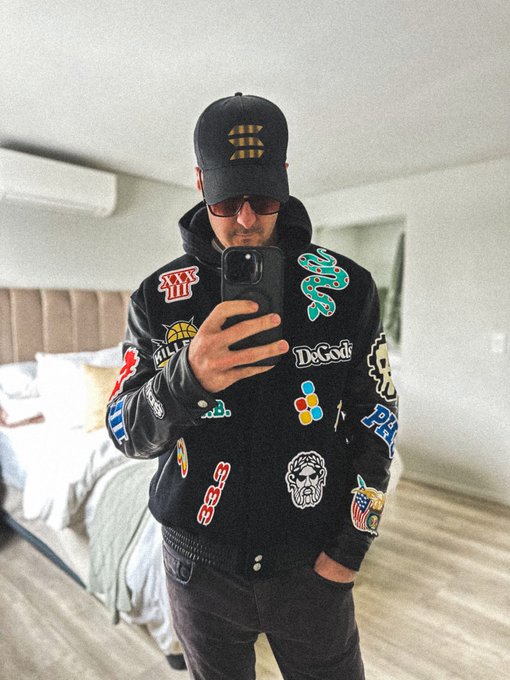 The image features a man wearing a black jacket with various stickers, including one that says "DOGO" and another that says "PHIL". He is also wearing sunglasses and holding up his phone for a selfie. The background of the photo shows a bedroom setting with a bed visible in the room. The post includes text such as "Woke up this morning and decided to be a walking billboard for @solana and @DeGodsNFT", which suggests that the man is promoting these brands or products through his selfie.