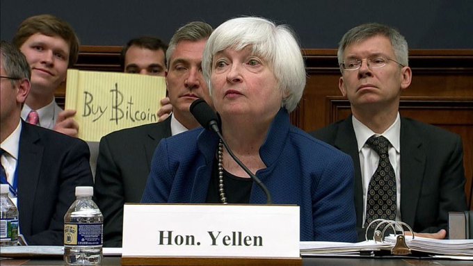 Former U.S. Treasury Secretary Janet Yellen, identified by the 'Hon. Yellen' nameplate, testifies at a congressional hearing while someone behind her holds a 'Buy Bitcoin' sign.