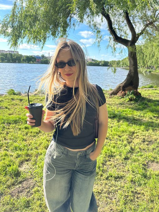 A blonde woman wearing sunglasses, a black crop top, and baggy jeans, holding a black cup and earphones while standing by a lake.