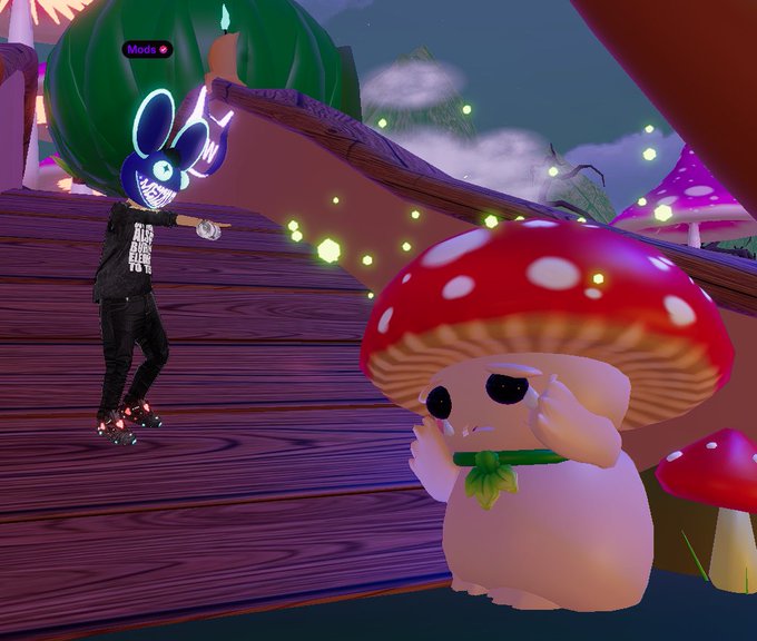 A player with a blue bunny mask and black clothing stands on a wooden platform in a virtual environment, with a "Mods" label visible above their head. Nearby, an adorable mushroom-like creature with a red spotted cap and small fangs poses with its hands raised, set against a fantastical backdrop with floating lights and oversized mushrooms.