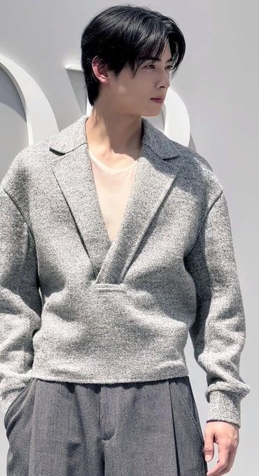 A young man with dark hair wearing a gray knit cardigan over a beige t-shirt stands against a plain white wall, looking off to the side.