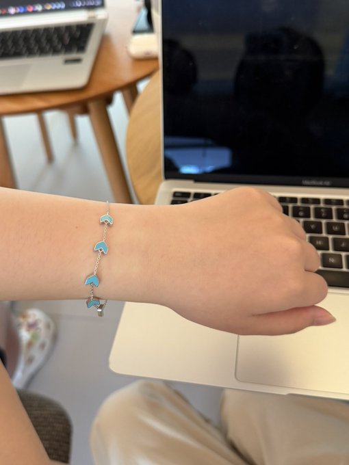 A close-up of someone's wrist wearing a delicate silver bracelet adorned with turquoise dolphin charms, resting near a MacBook Pro keyboard to showcase the fair skin tone and the rare Bitget Wallet merchandise piece.