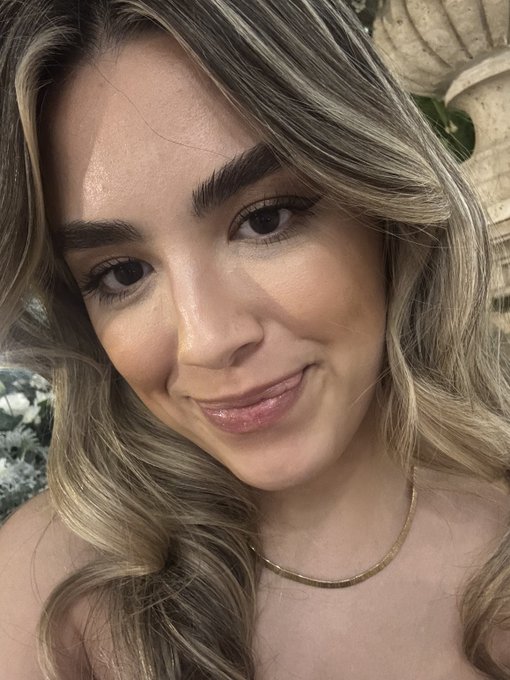 Close-up selfie of a woman with long blonde-brown hair wearing a gold necklace, with greenery and a stone architectural element visible in the background.