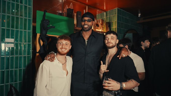 Three friends pose together at a lively nighttime venue, with the taller man in the center wearing a black cap, sunglasses, and a dark button-up shirt, while the man on the left wears a cream shirt and the man on the right flashes a peace sign while holding a drink, with a green "GM" logo visible on the wall behind them—tying into the crypto community's "GM" (good morning) greeting often used as a casual sign-off among Web3 enthusiasts.