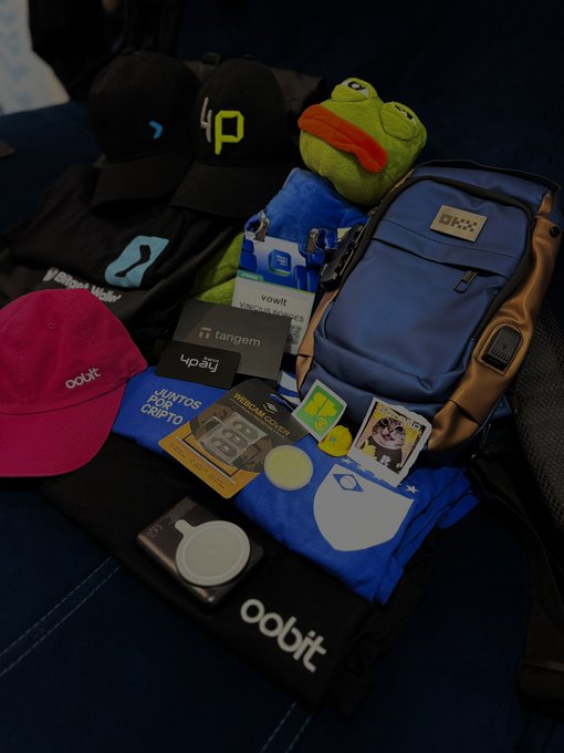 A collection of crypto and tech promotional swag, including hats for Oobit and other brands, a blue backpack, t-shirts, stickers, and a green Pepe the Frog plush toy.