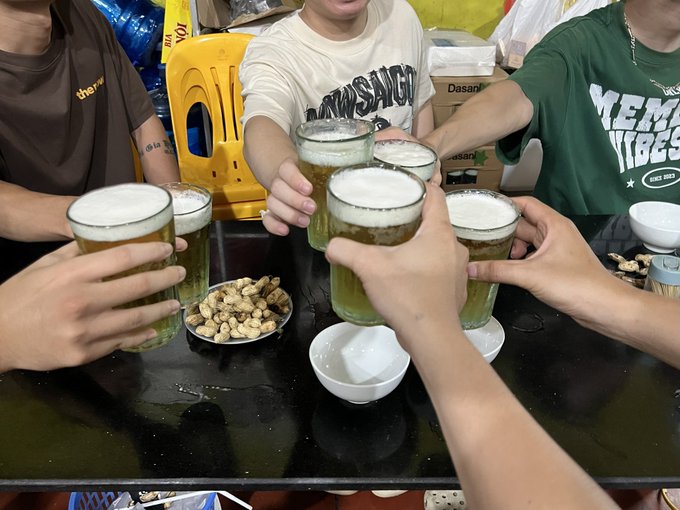 A group of friends clinks glasses of frothy beer together over a black table featuring a small plate of peanuts and a white bowl, in what appears to be a casual Asian eatery.