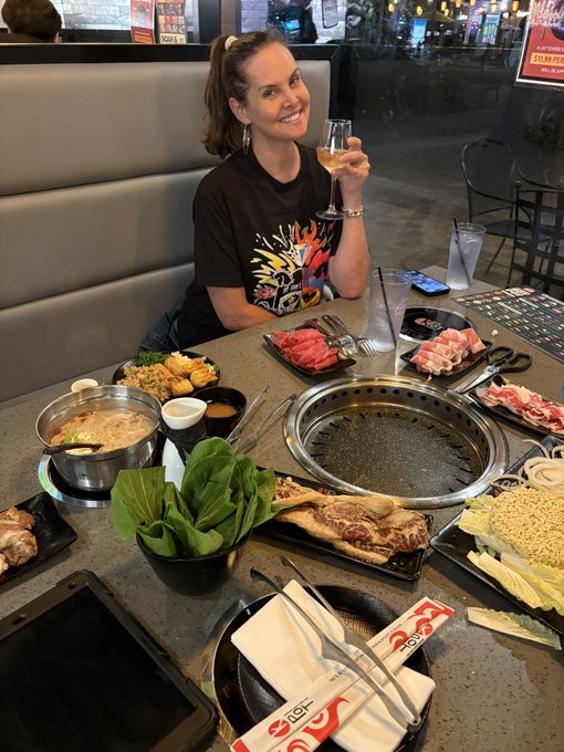 A smiling woman sits at a Korean BBQ restaurant table filled with plates of raw meat, vegetables, and a tabletop grill, holding up a wine glass.