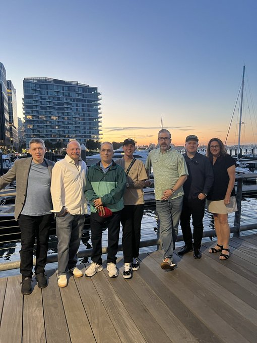 Seven people pose together on a wooden dock at a Boston marina during sunset, with sailboat masts and a waterfront building visible in the background. The caption's mention of "Good Company in Boston" and gathering people together aligns with the group photo, suggesting a social networking event where the team welcomed newcomers ("normies") into their community.