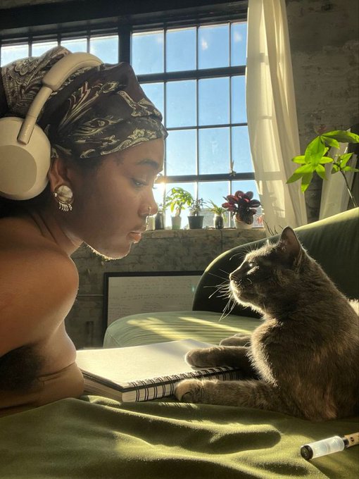 The image shows a woman wearing headphones sitting next to her cat on a couch. She has an open notebook or book placed beside her, suggesting that she might be studying or working on something related to AI. The caption of the image reads "Happy Monday CT frens It' s a new week again what will you be working on today? I'll be positioning on @quipnetwork & $RIVER And also studying the fact that @TheARCTERMINAL is making AI more productive, autonomous, private, secure, and user-controlled."