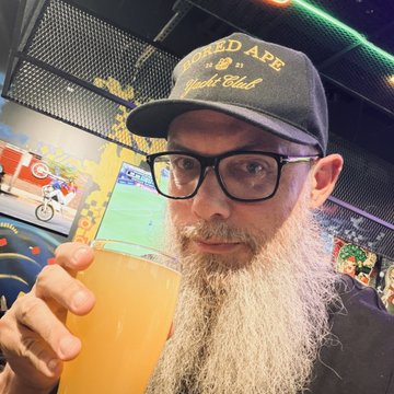 A bearded man wearing glasses and a "Funked Ape Yacht Club" cap holds up a hazy, golden-orange beer at Bronx Brewery, with a TV screen in the background showing a FIFA World Cup match. The image captures the moment he's enjoying the "Smile My Guy Happy Hoppy IPA," reflecting the post's reflection on how the familiar sip from ApeFest 2022 brings back memories.