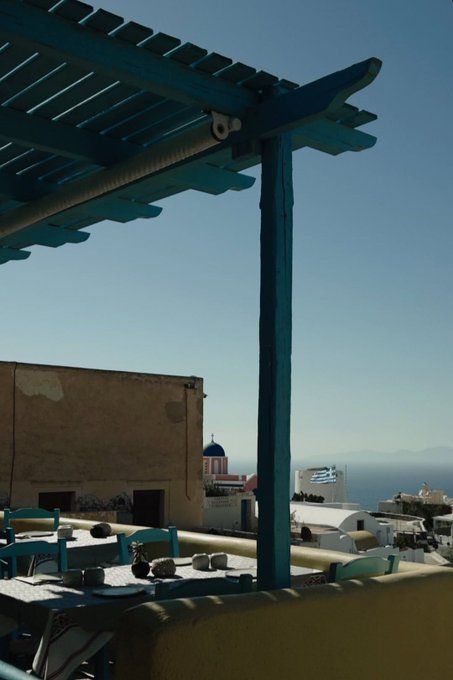 A rooftop terrace with a turquoise wooden pergola overlooks the sun-bleached whitewashed buildings and blue-domed church of Santorini, Greece, with the Aegean Sea shimmering in the distance—perfectly capturing the "Greecemaxxing" island escape.