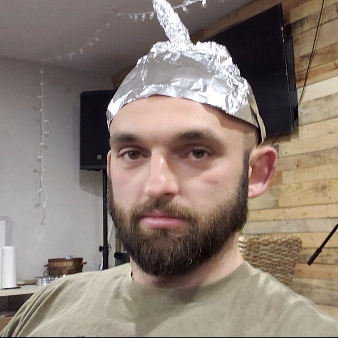 A bearded man wears a crumpled tinfoil hat shaped like a cone, suggesting an eccentric or "conspiracy theorist" aesthetic, which humorously matches his post about using vintage calculators and BIP-39 word lists for Bitcoin-related calculations.