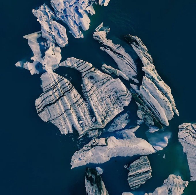 An aerial view of fragmented icebergs floating in deep blue water, with some pieces showing distinctive parallel striations and textured surfaces.