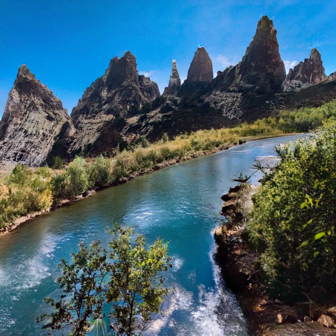A scenic landscape showing turquoise river waters flowing past jagged, rocky mountain peaks under a clear blue sky, with green vegetation along the riverbanks.