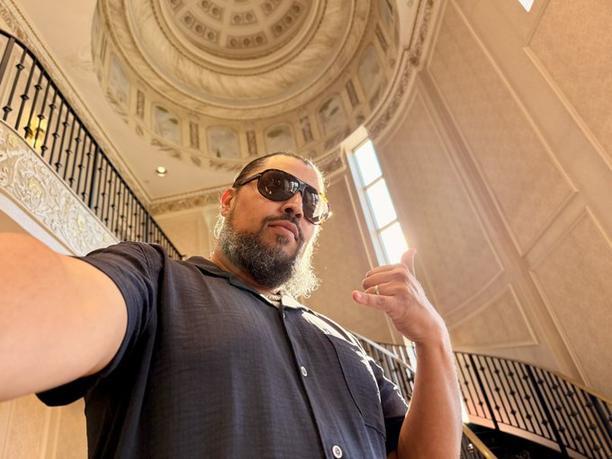 A bearded man in sunglasses takes a low-angle selfie in an ornate domed hall with elegant railings, while his caption "My voice is bigger than the NFT I wear. IYKYK" suggests his confident expression and raised finger gesture emphasize the bold statement about his voice outshining any wearable digital art.