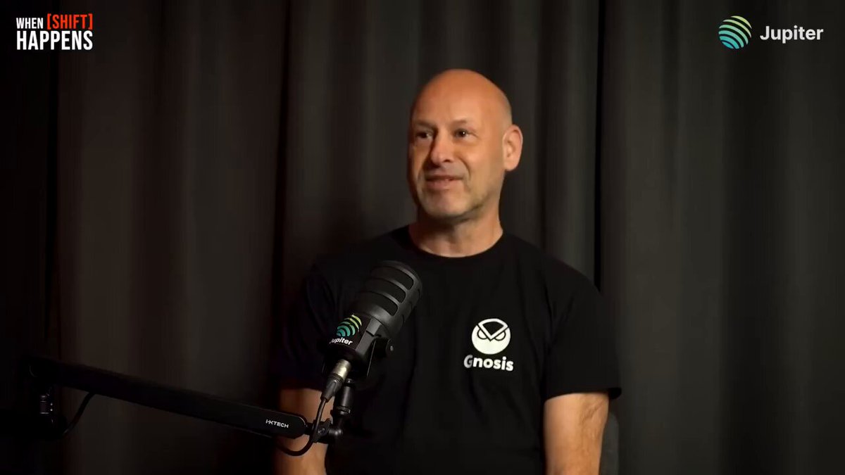 The image features an older man wearing a black t-shirt with white text, standing behind a microphone and speaking into it. The background shows a dark curtain or screen, which contrasts with the man's attire. The caption of the image reads "Focus on what is in the image and how it relates to the post".