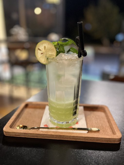 A tall glass of green cocktail garnished with a lime slice and fresh basil sits on a wooden tray with a bar spoon, served in a moody bar setting—likely a gin-based drink fitting the "Gn bros" (gin bros) caption.