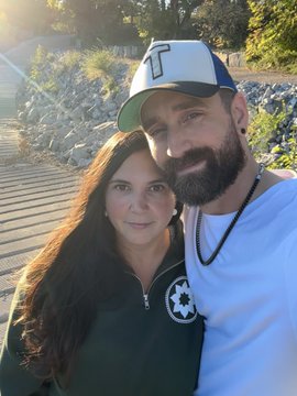 A couple taking a selfie outdoors; the man wears a baseball cap, white t-shirt, and beaded necklace, while the woman wears a dark green hoodie.