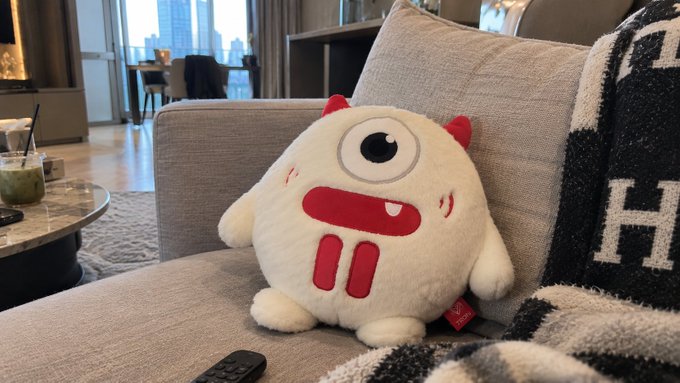 A white plush "TRON Bull" mascot with red horns, one large eye, and a wide smile sits on a gray sofa in a modern apartment, alongside a black and white blanket and a TV remote. The caption ties the image to TRON crypto community swag becoming a permanent fixture in the creator's daily life, with the plush symbolizing ongoing representation of the TRON brand at home.