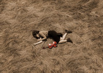 An aerial view shows a person in a black top and shorts lying on dry, golden grass, holding a small red chainsaw next to a long stick or pole.
