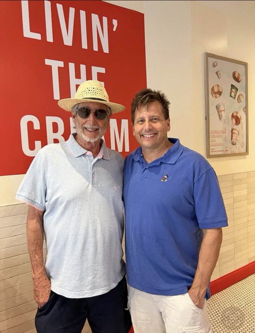 Two men pose together in front of a large red sign reading "LIVIN' THE CREAM" (with "CREAM" partially obscured), smiling warmly for a photo that captures a sweet father-son moment.