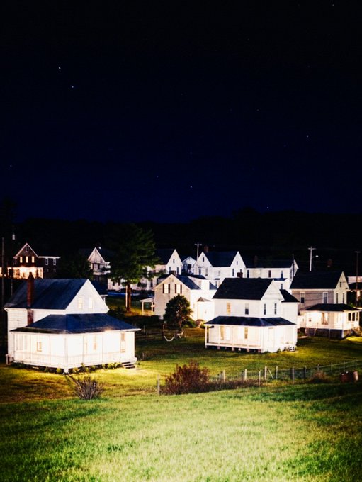 A nighttime scene shows a cluster of small white houses with lit windows, surrounded by green grass under a dark starry sky.