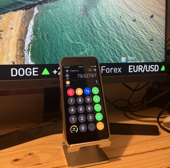 A smartphone displaying a CryptoCalc calculator app shows a Bitcoin-to-USD conversion of 79,527.67, propped in front of a monitor displaying a beach scene and a ticker for DOGE and EUR/USD.