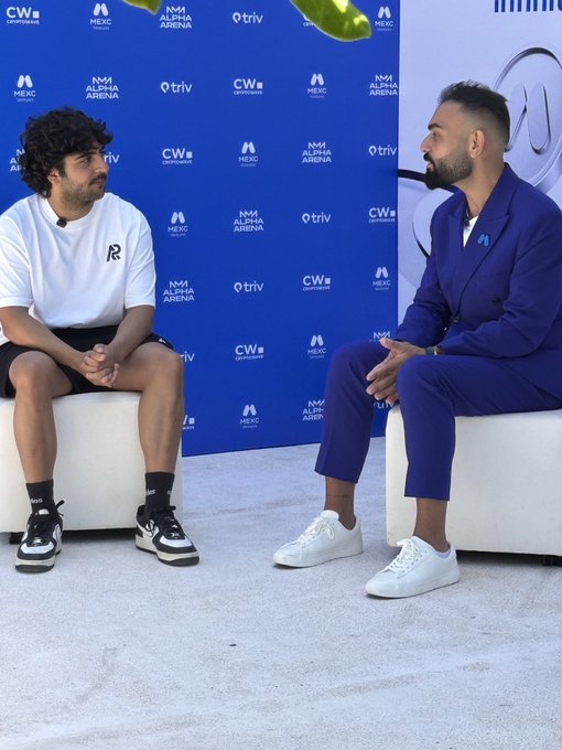 Two men seated on white chairs having a discussion in front of a blue backdrop with logos for NM ALPHA ARENA, MEXC, triv, and CW cryptocurrencies.
