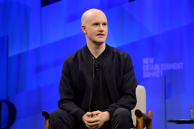 A bald man wearing a black bomber jacket sits in a chair on a stage with a blue backdrop that includes the text 'NEW ESTABLISHMENT SUMMIT'.