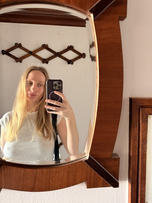 A mirror selfie of a blonde woman wearing a white top taking a photo with her smartphone, featuring a wooden mirror frame and a decorative wall rack in the background.