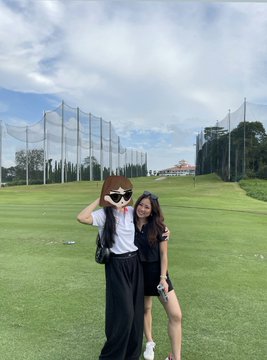 Two young women pose together outdoors on a grassy field, with tall netting structures visible in the background under a partly cloudy sky.