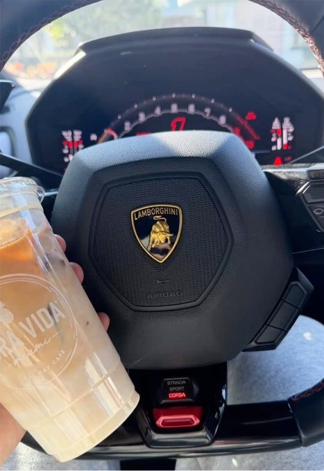 Interior view from the driver's seat of a Lamborghini with the steering wheel logo visible, a 'Strada/Sport/Corsa' mode selector, and a hand holding a 'Bravida' branded iced drink.