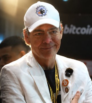 Max Keiser, wearing a hat with the logo "MAX IS A HACKER" and a Bitcoin-themed lanyard, smiles at the camera. The background prominently features the word "bitcoin," reinforcing the post's message.