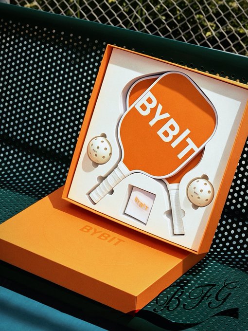 A Bybit-branded orange gift box containing paddle-shaped items and balls resembling a pickleball set, photographed on a metal mesh table with a watermark 'BFG'.