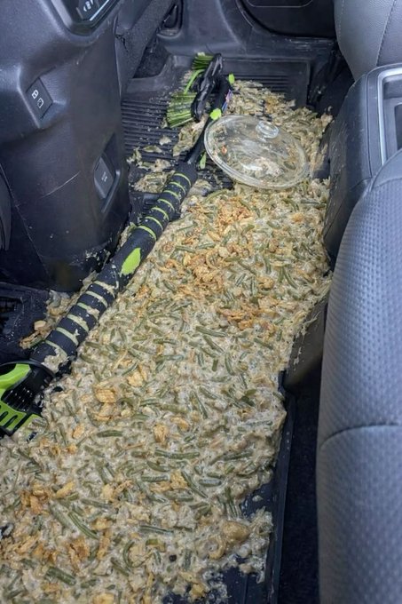 A casserole dish of green bean casserole has been completely crushed and spilled across the passenger floor of a Ford vehicle, leaving behind a messy carpet of beans, sauce, and toppings. The caption "What happened here?!!!" humorously highlights the disaster of transporting the holiday dish in the car.